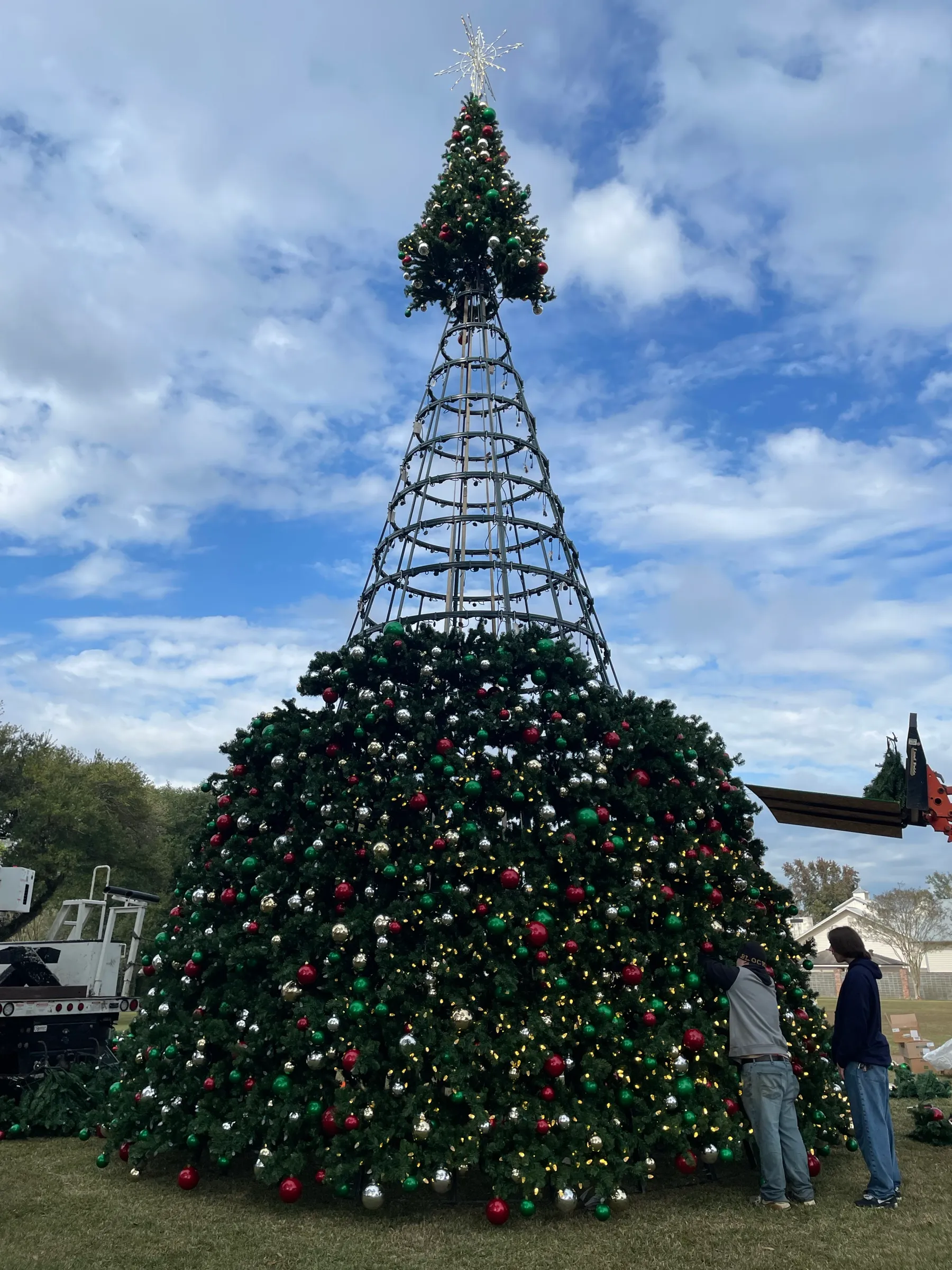Crew installing ornaments and lights on a large commercial Christmas tree during the day.