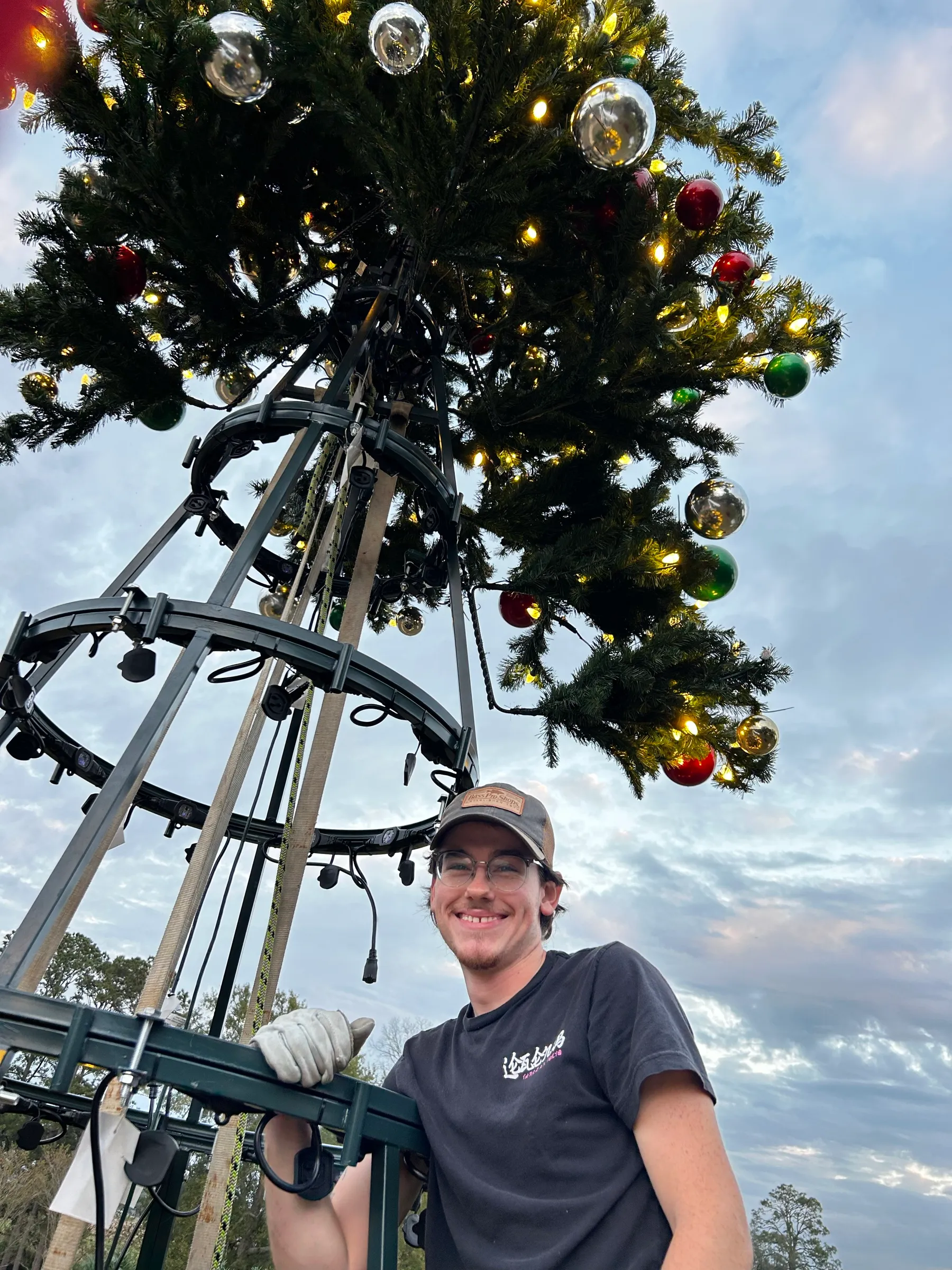 A crew member working on the steel frame of a commercial Christmas tree at height, attaching lit branch sections.