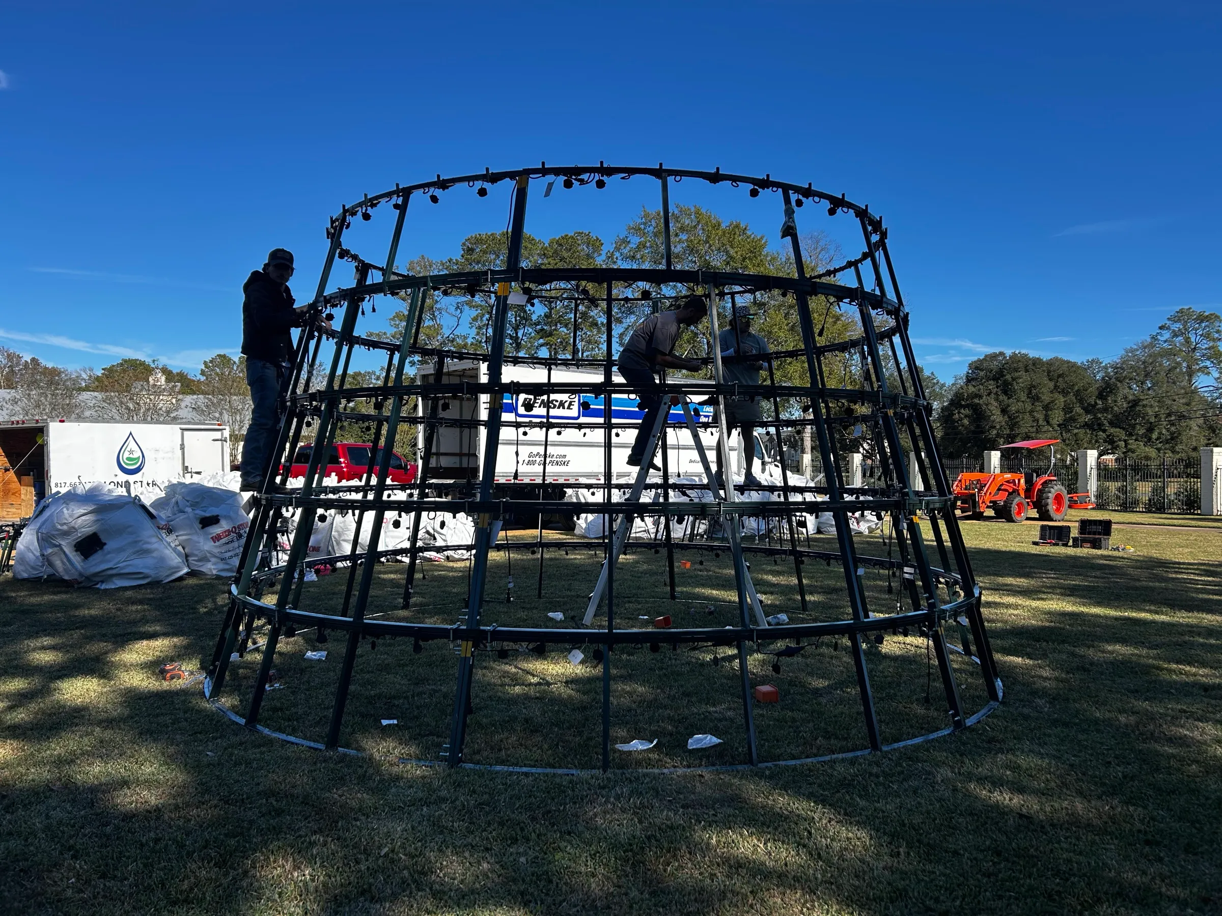 The steel ring frame of a commercial Christmas tree part way through assembly on a lawn in daylight.
